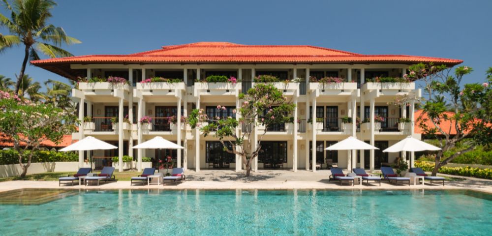 Three-story resort with red-tiled roof, balconies, and white umbrellas. A clear blue pool in the foreground, palm trees, and bright sky create a tranquil atmosphere.