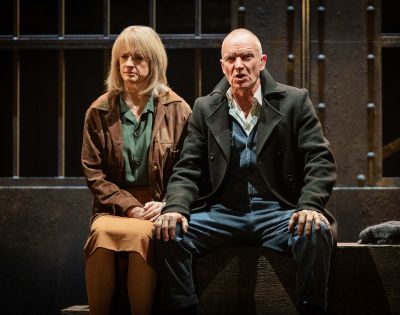 A man and a woman seated on a stage, both with serious expressions. They're dressed in earth-toned clothing, sitting in front of a backdrop resembling bars.