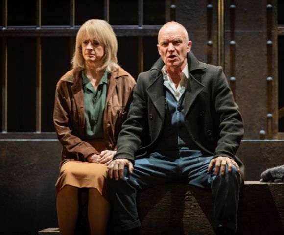 A man and a woman seated on a stage, both with serious expressions. They're dressed in earth-toned clothing, sitting in front of a backdrop resembling bars.