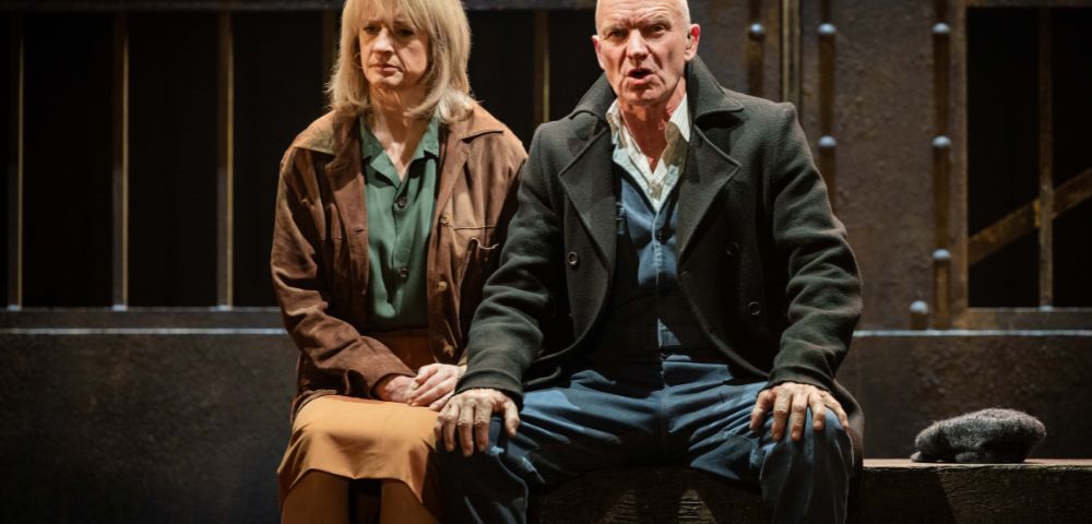 A man and a woman seated on a stage, both with serious expressions. They're dressed in earth-toned clothing, sitting in front of a backdrop resembling bars.