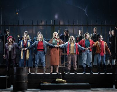 A group of performers stands on stage, linked arm in arm, passionately singing. They wear 20th-century casual attire, with a dramatic, intense backdrop.