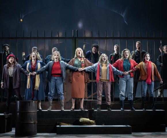 A group of performers stands on stage, linked arm in arm, passionately singing. They wear 20th-century casual attire, with a dramatic, intense backdrop.