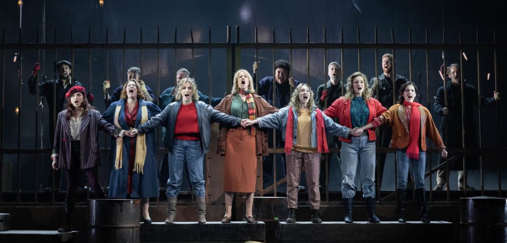 A group of performers stands on stage, linked arm in arm, passionately singing. They wear 20th-century casual attire, with a dramatic, intense backdrop.