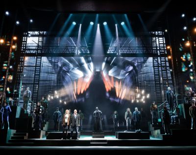 A dramatic stage scene with a large cast and dynamic lighting. Central actors stand under spotlights; a giant hand graphic dominates the backdrop.