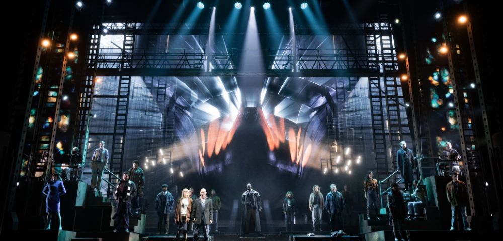 A dramatic stage scene with a large cast and dynamic lighting. Central actors stand under spotlights; a giant hand graphic dominates the backdrop.