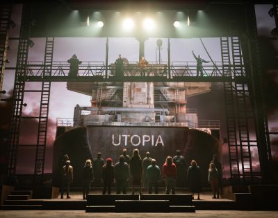 A dramatic stage scene depicts people standing on and around a large ship with 
