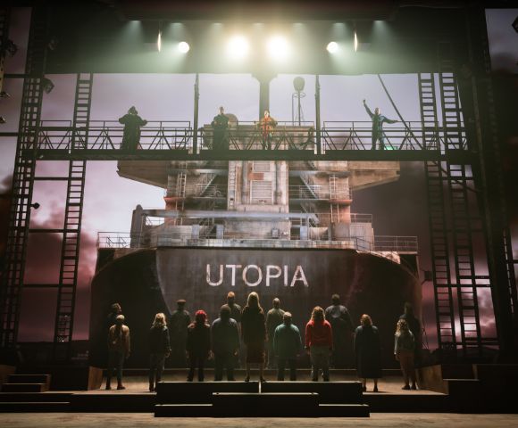 A dramatic stage scene depicts people standing on and around a large ship with 