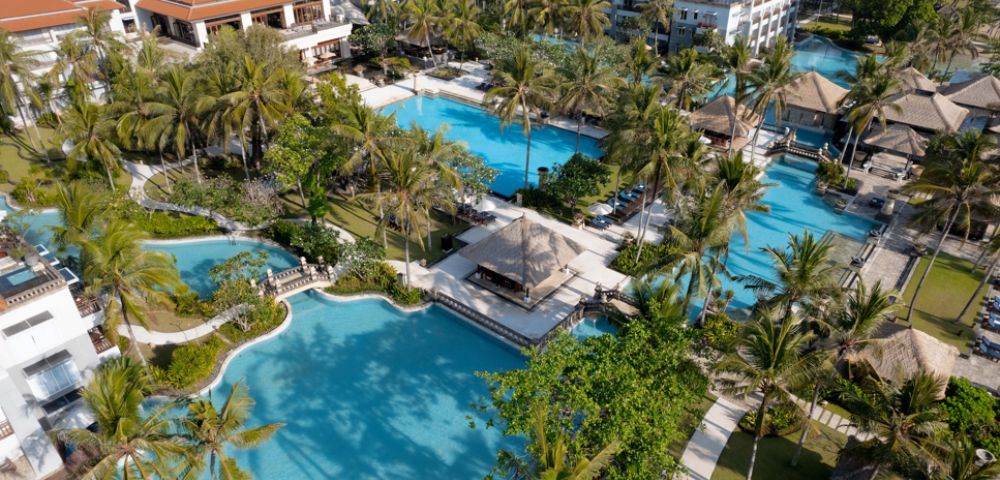 Aerial view of a tropical resort featuring turquoise pools winding through lush green palm trees, with red-roofed buildings in the background, exuding relaxation.