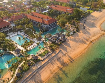 Aerial view of a beachfront resort with red-roofed buildings, multiple pools, and palm trees. The sandy beach meets clear turquoise water, casting long shadows.