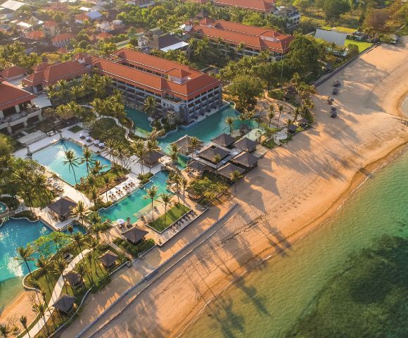Aerial view of a beachfront resort with red-roofed buildings, multiple pools, and palm trees. The sandy beach meets clear turquoise water, casting long shadows.