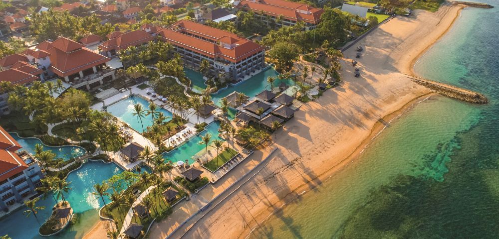 Aerial view of a beachfront resort with red-roofed buildings, multiple pools, and palm trees. The sandy beach meets clear turquoise water, casting long shadows.