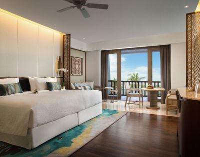 Luxurious hotel room with a large bed, elegant decor, and a colorful rug. A balcony with wooden chairs offers a view of palm trees and a blue sky.