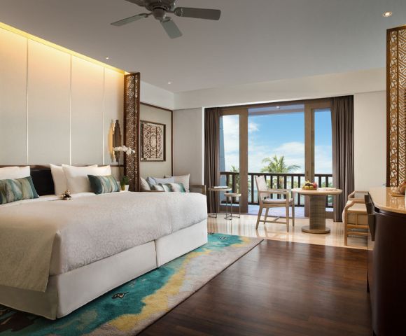 Luxurious hotel room with a large bed, elegant decor, and a colorful rug. A balcony with wooden chairs offers a view of palm trees and a blue sky.