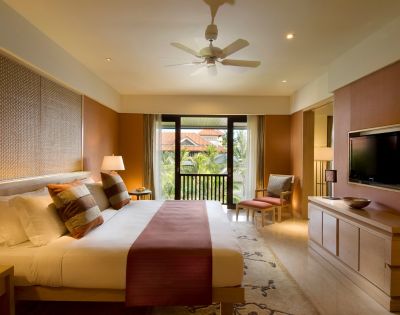 Modern hotel room with a large bed, patterned cushions, and a burgundy throw. A ceiling fan spins above. There's a flat-screen TV and a balcony view.