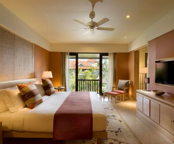 Modern hotel room with a large bed, patterned cushions, and a burgundy throw. A ceiling fan spins above. There's a flat-screen TV and a balcony view.