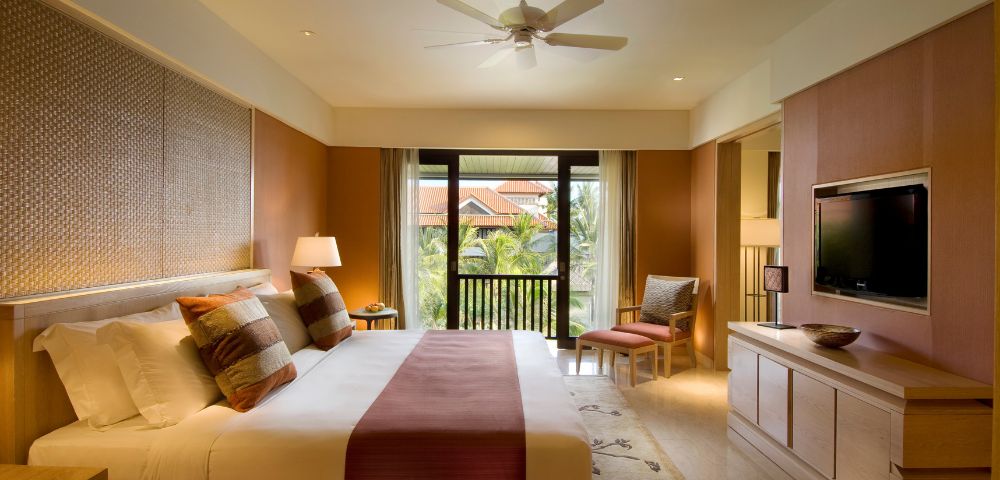 Modern hotel room with a large bed, patterned cushions, and a burgundy throw. A ceiling fan spins above. There's a flat-screen TV and a balcony view.