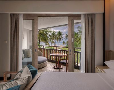 Elegant hotel room with open sliding doors leading to a balcony. The view reveals palm trees and a cloudy sky, creating a serene and inviting atmosphere.