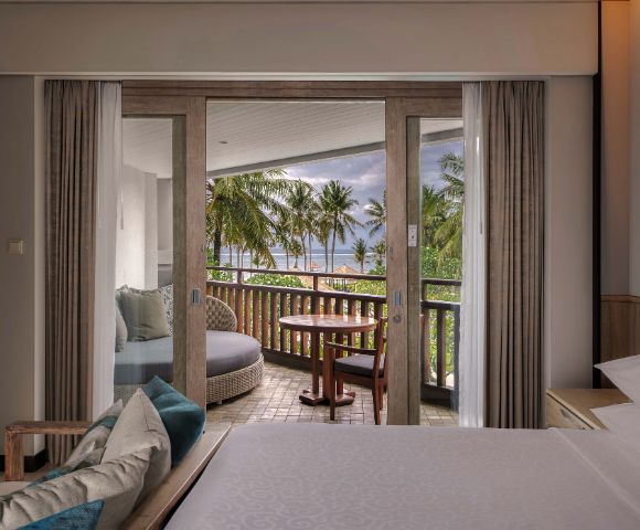 Elegant hotel room with open sliding doors leading to a balcony. The view reveals palm trees and a cloudy sky, creating a serene and inviting atmosphere.