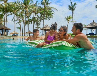 A family enjoying a sunny day in a tropical resort pool. Parents and two kids on a float laugh and play, surrounded by palm trees and cabanas.