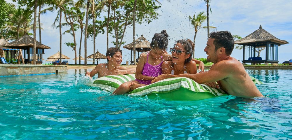 A family enjoying a sunny day in a tropical resort pool. Parents and two kids on a float laugh and play, surrounded by palm trees and cabanas.