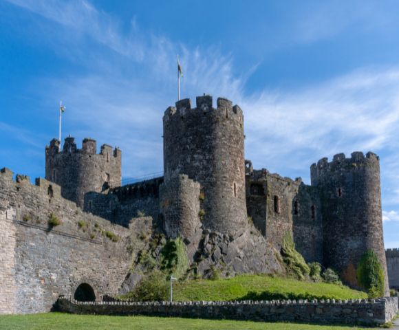 Ancient stone castle with round towers and battlements under a vibrant blue sky. Flags atop the towers add a sense of history and grandeur.