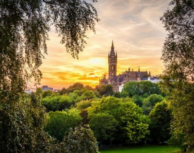Sunset view of a historic Gothic-style building framed by lush green trees. The sky is vibrant with orange and pink hues, creating a peaceful, romantic ambiance.