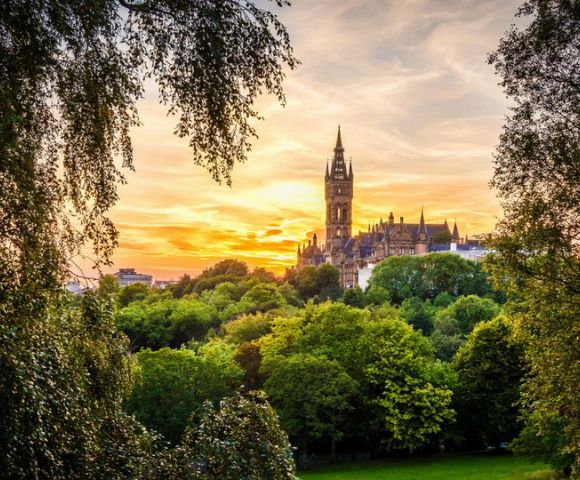 Sunset view of a historic Gothic-style building framed by lush green trees. The sky is vibrant with orange and pink hues, creating a peaceful, romantic ambiance.