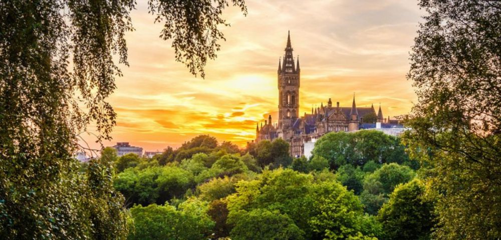 Sunset view of a historic Gothic-style building framed by lush green trees. The sky is vibrant with orange and pink hues, creating a peaceful, romantic ambiance.