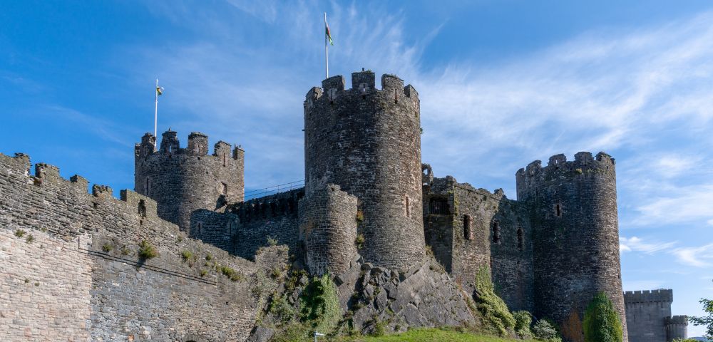 Majestic medieval castle under a clear blue sky. The stone fortress features round towers, battlements, and flags, exuding historic grandeur and strength.