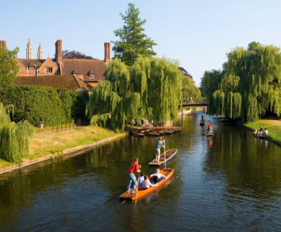 People are punting on a calm river lined with lush willow trees. A historic brick building is in the background under a clear blue sky. Peaceful atmosphere.