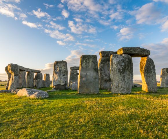 An ancient stone circle of large rocks under a blue sky with clouds, bathed in warm sunset light. The scene conveys a sense of mystery and timelessness.