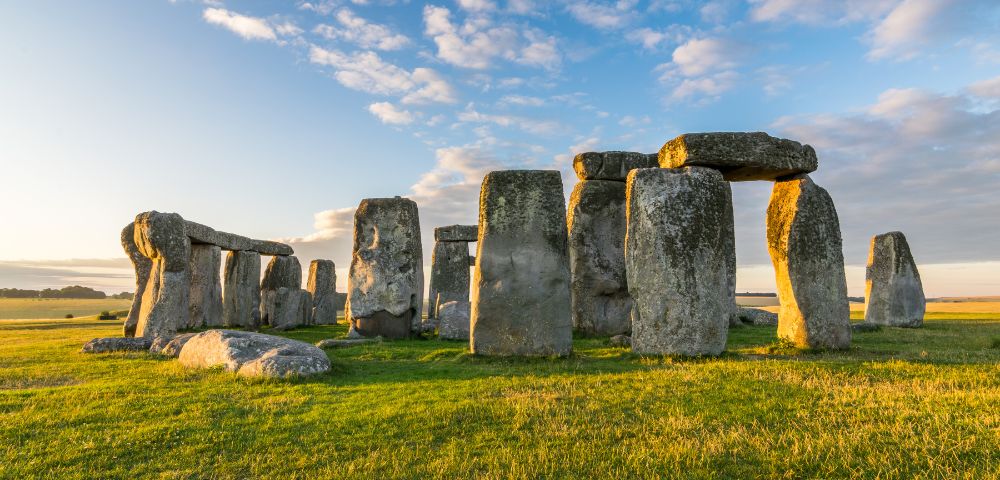 An ancient stone circle of large rocks under a blue sky with clouds, bathed in warm sunset light. The scene conveys a sense of mystery and timelessness.