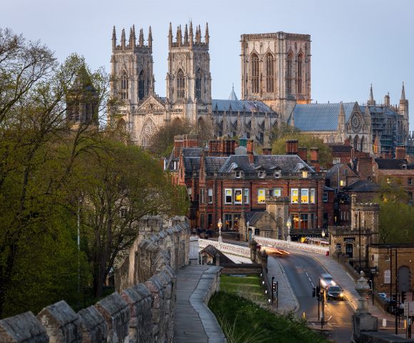 York Minster towers over a historic cityscape at dusk, with a stone wall and trees in the foreground and car lights illuminating the street below.