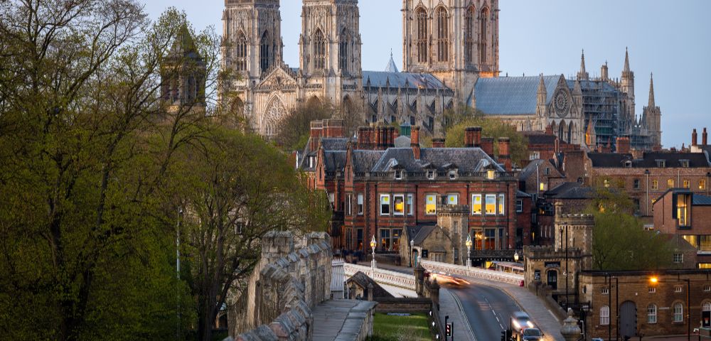 York Minster towers over a historic cityscape at dusk, with a stone wall and trees in the foreground and car lights illuminating the street below.