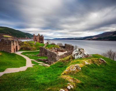 Ancient stone ruins on lush green hills overlook a vast lake under a dramatic, cloudy sky. A winding path leads through the tranquil landscape.