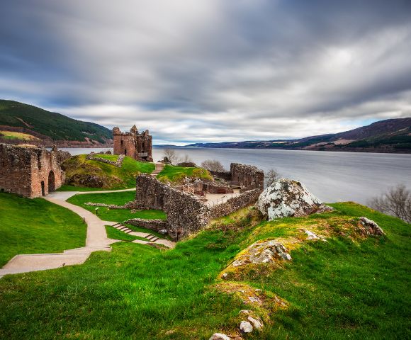 Ancient stone ruins on lush green hills overlook a vast lake under a dramatic, cloudy sky. A winding path leads through the tranquil landscape.