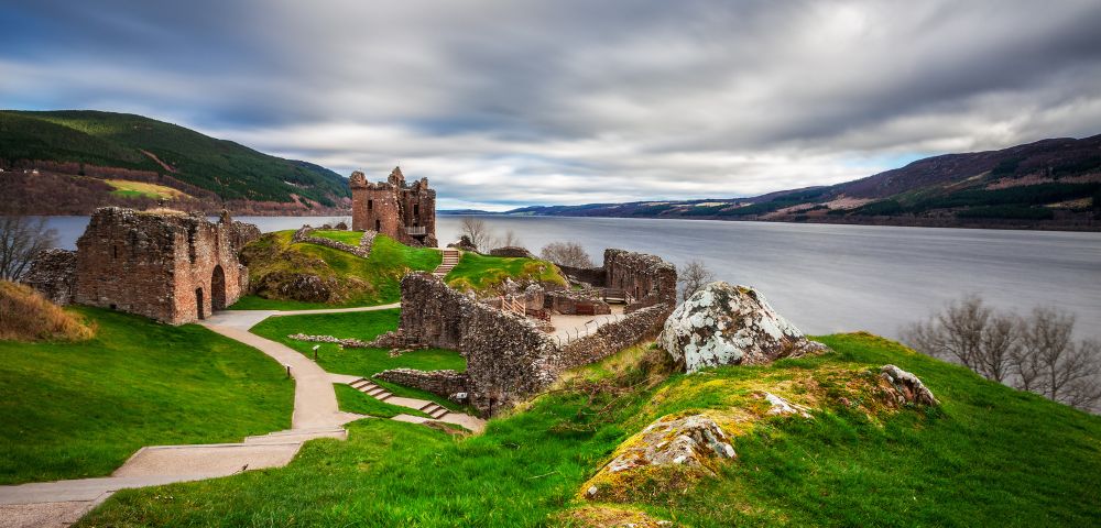 Ancient stone ruins on lush green hills overlook a vast lake under a dramatic, cloudy sky. A winding path leads through the tranquil landscape.