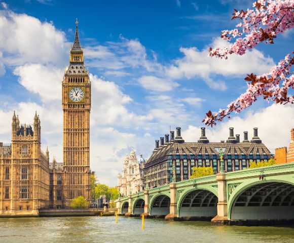 View of Big Ben and the Houses of Parliament in London on a sunny day with a clear blue sky. Cherry blossoms frame the scene in the foreground.