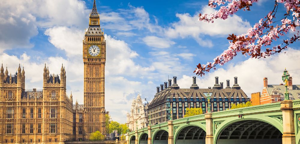 View of Big Ben and the Houses of Parliament in London on a sunny day with a clear blue sky. Cherry blossoms frame the scene in the foreground.