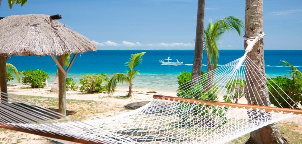 A hammock tied between palm trees on a sandy beach overlooks a calm turquoise sea. A thatched umbrella and a small boat enhance the serene tropical setting.