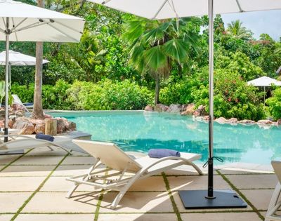 Poolside scene with white lounge chairs and umbrellas on a stone patio, surrounded by lush green trees, evoking a serene and tropical ambiance.