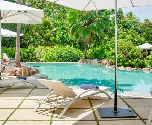 Poolside scene with white lounge chairs and umbrellas on a stone patio, surrounded by lush green trees, evoking a serene and tropical ambiance.