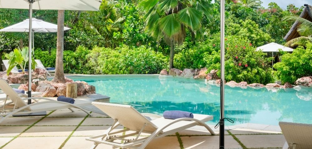 Poolside scene with white lounge chairs and umbrellas on a stone patio, surrounded by lush green trees, evoking a serene and tropical ambiance.
