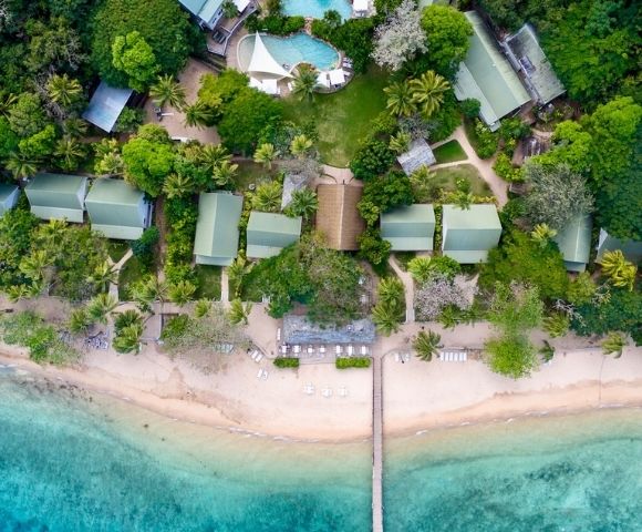 Aerial view of a tropical beach with turquoise water, lined with green-roofed buildings surrounded by lush trees, a wooden pier, and white sand.