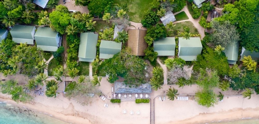 Aerial view of a tropical beach with turquoise water, lined with green-roofed buildings surrounded by lush trees, a wooden pier, and white sand.