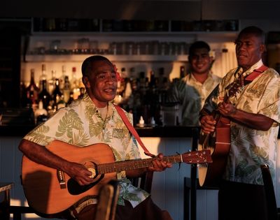 Three men in Hawaiian shirts play guitars and sing at a dimly lit bar, creating a relaxed, tropical ambiance with visible liquor bottles behind.