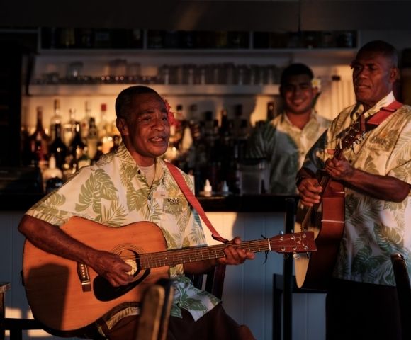 Three men in Hawaiian shirts play guitars and sing at a dimly lit bar, creating a relaxed, tropical ambiance with visible liquor bottles behind.