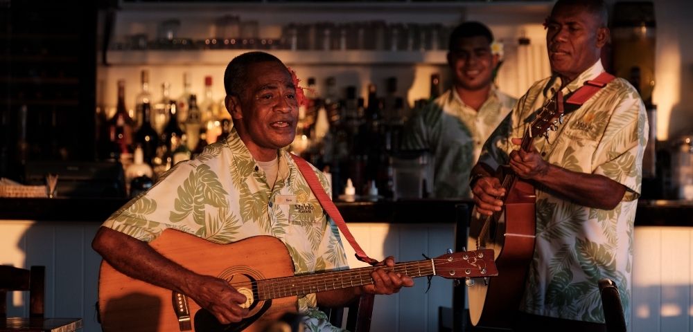 Three men in Hawaiian shirts play guitars and sing at a dimly lit bar, creating a relaxed, tropical ambiance with visible liquor bottles behind.