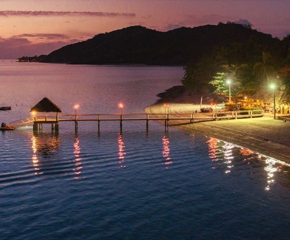 Tranquil beach scene at sunset with a wooden pier extending into calm water. Soft, warm lights illuminate the pier and reflect on the ocean.