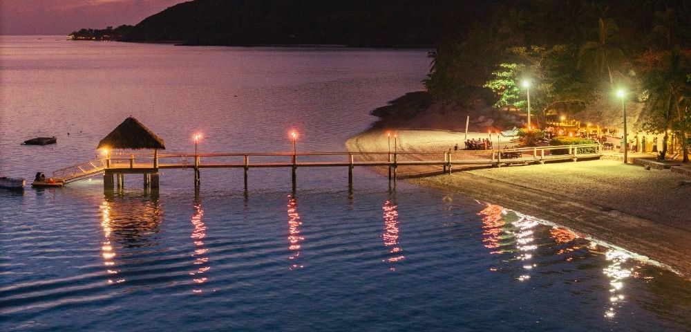 Tranquil beach scene at sunset with a wooden pier extending into calm water. Soft, warm lights illuminate the pier and reflect on the ocean.
